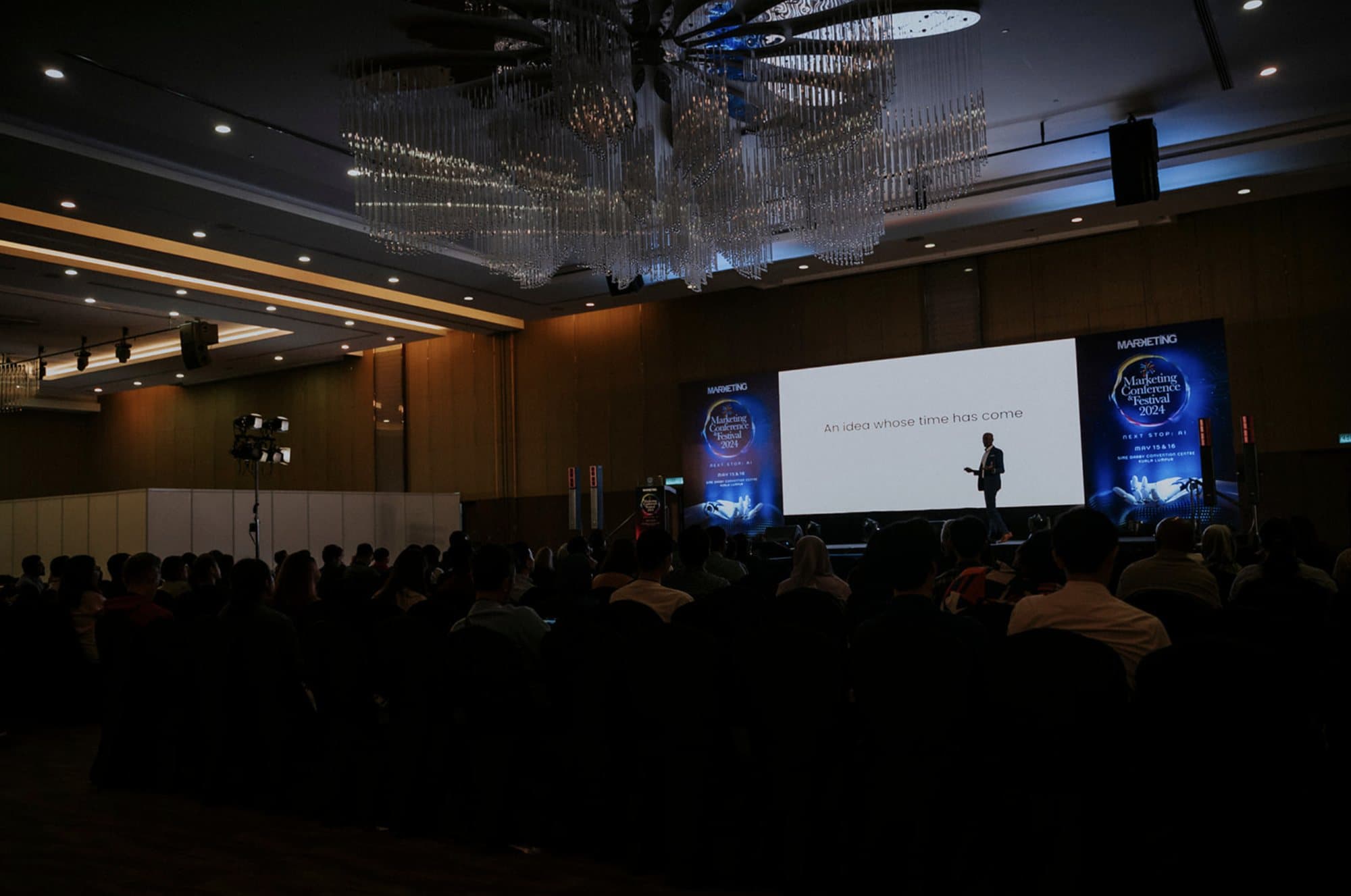 A full conference hall of seated delegates facing a lit stage where Indhran Seghar is speaking